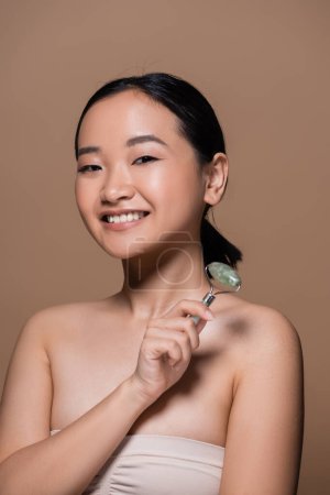 Foto de Positive asian woman in top holding jade roller and looking at camera isolated on brown - Imagen libre de derechos