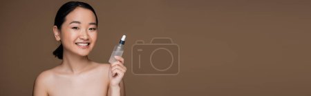 Smiling asian model with naked shoulders holding cosmetic serum isolated on brown, banner 
