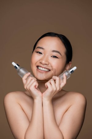 Photo for Cheerful asian woman with naked shoulders holding cosmetic serums isolated on brown - Royalty Free Image
