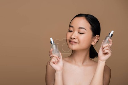 Photo for Pretty asian woman with naked shoulders holding bottles of serums isolated on brown - Royalty Free Image