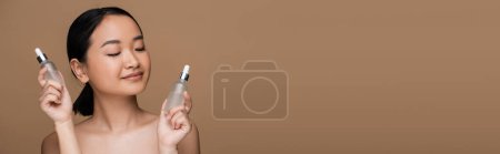 Brunette asian woman with naked shoulders holding bottles of cosmetic serums isolated on brown, banner 