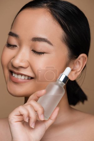 Close up view of smiling asian model with closed eyes holding serum isolated on brown 