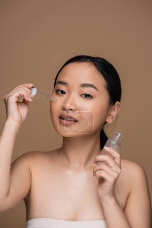 Brunette asian woman applying serum and looking at camera isolated on brown 