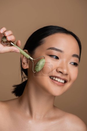 Positive asian model with naked shoulders using jade roller on face isolated on brown 