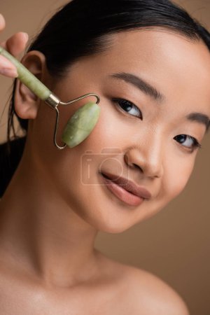 Téléchargez les photos : Portrait of brunette asian woman using jade roller on face isolated on brown - en image libre de droit