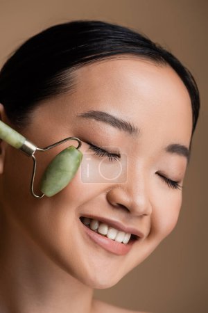 Téléchargez les photos : Pleased asian woman using jade roller on cheek isolated on brown - en image libre de droit