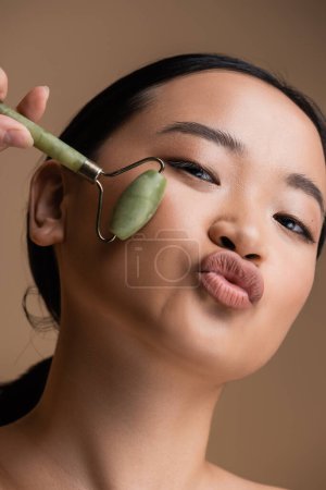 Foto de Portrait of asian woman holding jade roller and pouting lips isolated on brown - Imagen libre de derechos