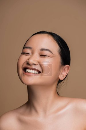 Cheerful asian woman with visage and naked shoulders standing isolated on brown 