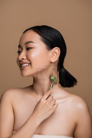 Photo for Happy asian woman with naked shoulders using jade roller on neck isolated on brown - Royalty Free Image