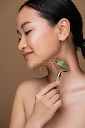 Asian woman with naked shoulder using jade roller on neck isolated on brown 
