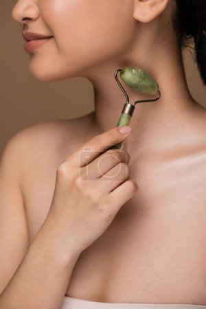 Cropped view of young woman holding jade roller near neck isolated on brown 