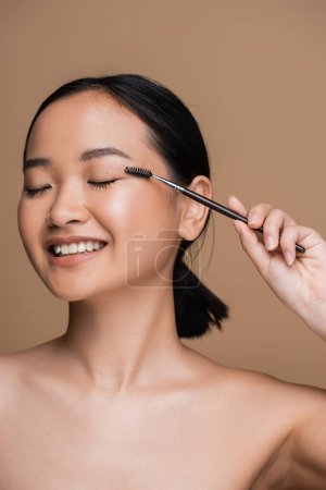 Positive asian woman with naked shoulders holding eyebrow brush isolated on brown 