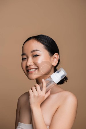 Young asian woman with naked shoulders holding face cleanser isolated on brown 