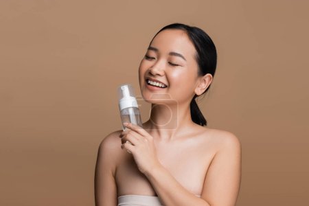 Positive asian model with naked shoulders holding cleansing foam isolated on brown 