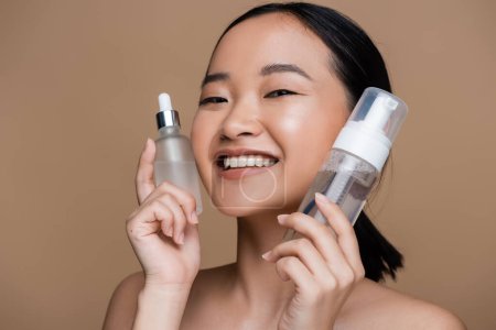 Positive asian woman with naked shoulders holding serum and face cleanser isolated on brown 