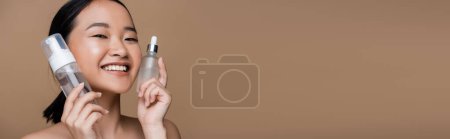 Téléchargez les photos : Cheerful asian woman holding face cleanser and serum isolated on brown, banner - en image libre de droit