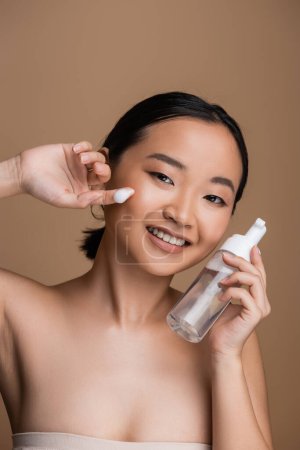 Positive asian model with naked shoulders holding cleanser with dispenser isolated on brown 