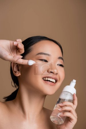 Cheerful asian woman with naked shoulders holding bottle of face foam isolated on brown 