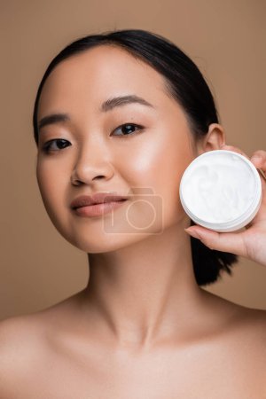 Téléchargez les photos : Pretty asian woman with naked shoulders and perfect skin holding cosmetic cream isolated on brown - en image libre de droit