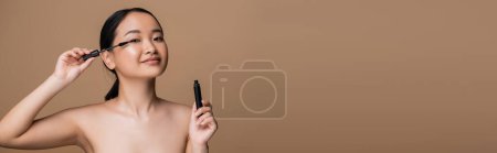Foto de Pretty asian woman with naked shoulders holding mascara isolated on brown, banner - Imagen libre de derechos