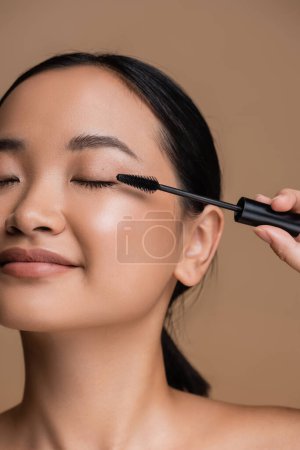 Foto de Cropped view of young asian woman holding applicator for mascara isolated on brown - Imagen libre de derechos