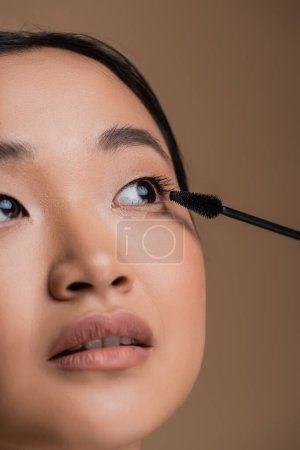 Foto de Cropped view of asian woman with makeup holding mascara applicator isolated on brown - Imagen libre de derechos