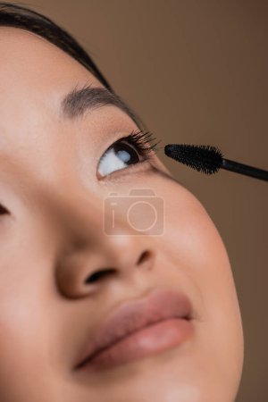 Foto de Close up view of asian woman applying mascara isolated on brown - Imagen libre de derechos