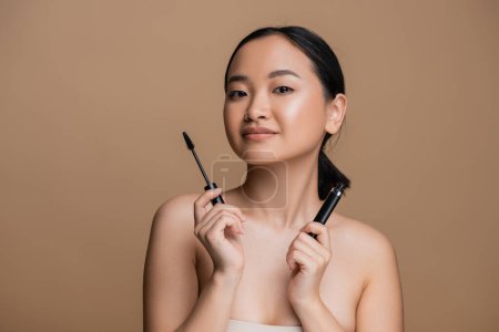 Foto de Pretty asian woman holding mascara and looking at camera isolated on brown - Imagen libre de derechos