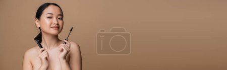 Téléchargez les photos : Young asian woman with naked shoulders holding mascara isolated on brown, banner - en image libre de droit