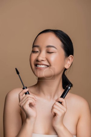 Foto de Cheerful asian woman with naked shoulders holding mascara isolated on brown - Imagen libre de derechos