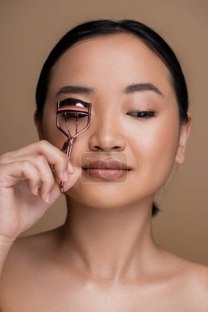 Foto de Asian model with makeup using lush curler isolated on brown - Imagen libre de derechos