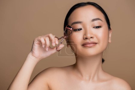 Pretty asian woman holding lush curler near face isolated on brown 