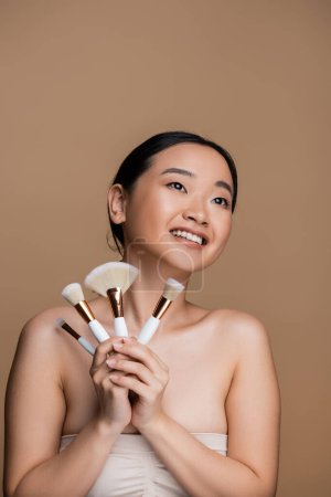 Photo for Positive asian model in top holding cosmetic brushes isolated on brown - Royalty Free Image