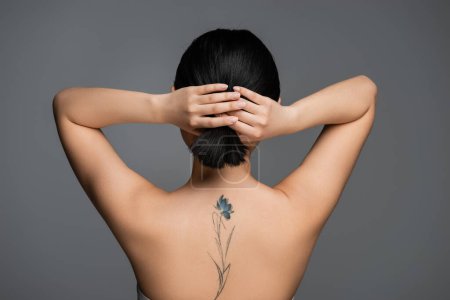 Back view of tattooed woman with naked shoulders touching ponytail isolated on grey 