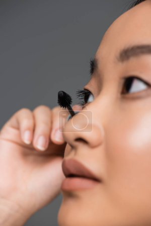 close up view of young asian woman applying black mascara isolated on grey