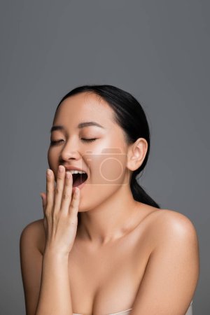 young asian woman with perfect skin and bare shoulders covering mouth with hand while yawning with closed eyes isolated on grey