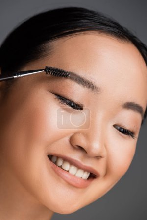 portrait of cheerful asian woman with natural makeup brushing eyebrow isolated on grey