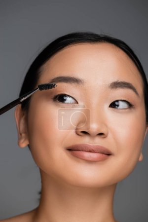 portrait of young asian woman with perfect skin brushing eyebrow isolated on grey