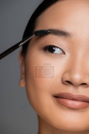 close up view of cropped asian woman with makeup foundation on face brushing eyebrow isolated on grey