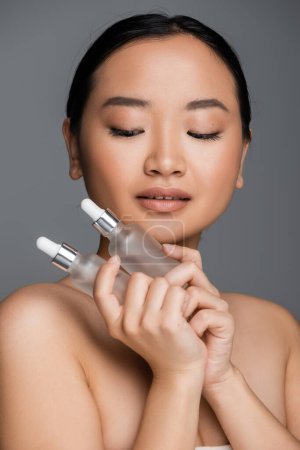 young asian woman with bare shoulders holding bottles with cosmetic serum isolated on grey