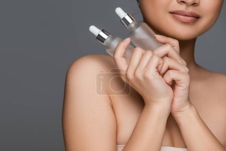 partial view of woman with perfect skin holding bottles with cosmetic serum isolated on grey