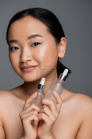 pleased asian woman with naked shoulders holding bottles with cosmetic serum and looking at camera isolated on grey