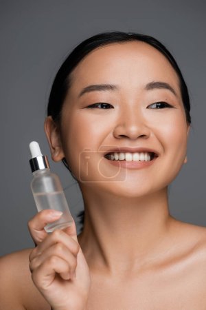 young and happy asian woman with clean skin holding bottle of cosmetic serum isolated on grey