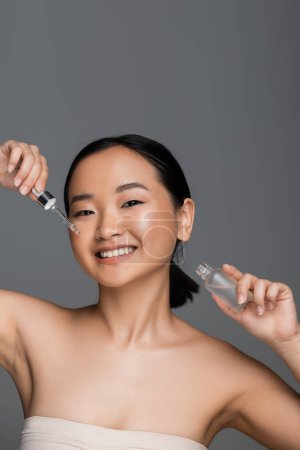 young asian woman holding bottle and dropper of cosmetic serum while smiling at camera isolated on grey