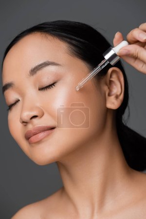 Photo for Young asian woman with closed eyes applying cosmetic serum on clean face isolated on grey - Royalty Free Image