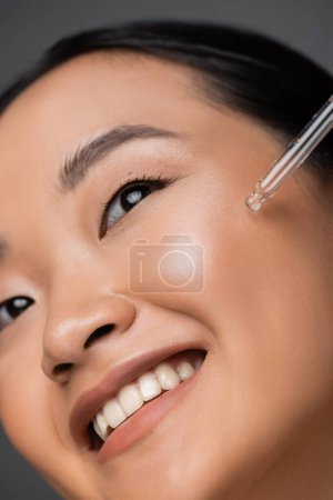 close up view of smiling asian woman applying cosmetic serum during facial pampering isolated on grey