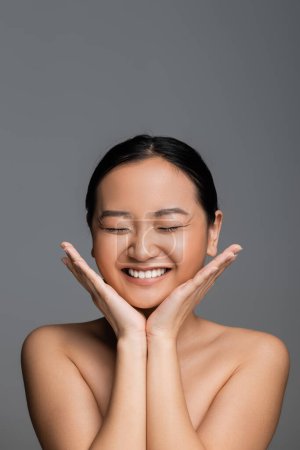 excited asian woman with closed eyes and nude makeup holding hands near face isolated on grey