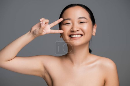 cheerful asian woman showing victory sign near face with natural makeup isolated on grey