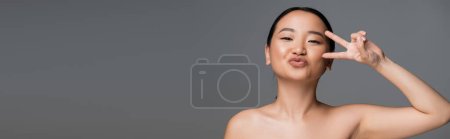 Foto de Young asian woman with bare shoulders showing victory sign and pouting lips isolated on grey, banner - Imagen libre de derechos