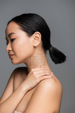 charming asian woman with nude makeup and ponytail hairstyle posing with hand on bare shoulder isolated on grey